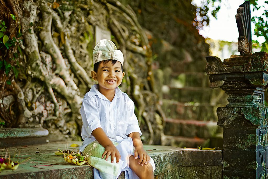 Die Versorgung im Krankheitsfall ist gewährleistet. Ein lachendes Kind sitzt neben einem Baum in tradtitioneller balinesischer Kleidung
